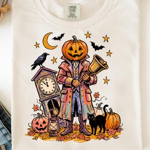 May include: A cream-colored t-shirt featuring a graphic of a pumpkin-headed figure in a long coat holding a bell. The design includes a grandfather clock, a black cat, a lantern, a raven, bats, stars, a crescent moon, and scattered autumn leaves.