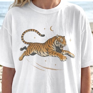 Puede incluir: Camiseta blanca con una ilustración de un tigre corriendo en tonos naranja y marrón. El diseño incluye una luna creciente y estrellas, con líneas curvas que sugieren movimiento. La camiseta está hecha de un material suave y cómodo.