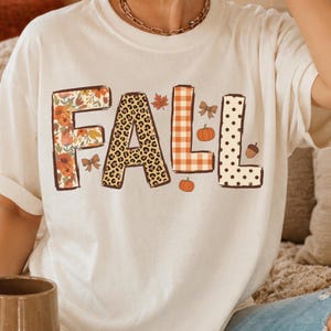 May include: A cream-colored short-sleeve t-shirt featuring the word FALL in large block letters. Each letter has a unique pattern: floral, leopard print, orange and white gingham, and polka dots, accented with small pumpkins, leaves, and bows.