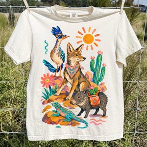 May include: A cream-colored short-sleeve t-shirt featuring a colorful graphic print of a roadrunner wearing sunglasses, a coyote with a patterned bandana, a javelina with a saddlebag, a lizard, a cactus, and a bright orange sun.