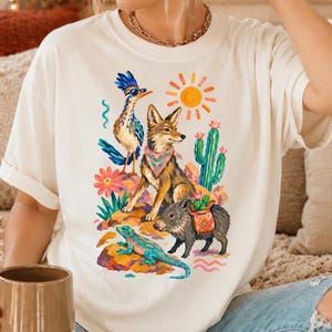 May include: An off-white t-shirt featuring a colorful graphic print of a desert scene. The illustration includes a roadrunner wearing sunglasses, a coyote with a patterned bandana, a javelina, a lizard, a cactus, and a bright orange sun.