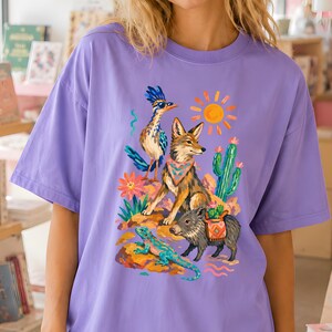 May include: A light purple t-shirt featuring a colorful graphic print of a desert scene. The illustration includes a roadrunner wearing sunglasses, a coyote with a patterned bandana, a javelina, a lizard, a cactus, and a bright yellow sun.