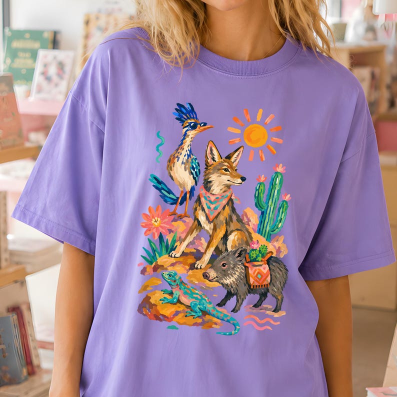 May include: A light purple t-shirt featuring a colorful graphic print of a desert scene. The illustration includes a roadrunner wearing sunglasses, a coyote with a patterned bandana, a javelina, a lizard, a cactus, and a bright yellow sun.