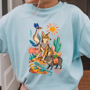 May include: A light blue crewneck sweatshirt featuring a colorful graphic print of a desert scene. The illustration includes a roadrunner wearing sunglasses, a coyote with a patterned bandana, a javelina, a lizard, a cactus, and a bright sun.