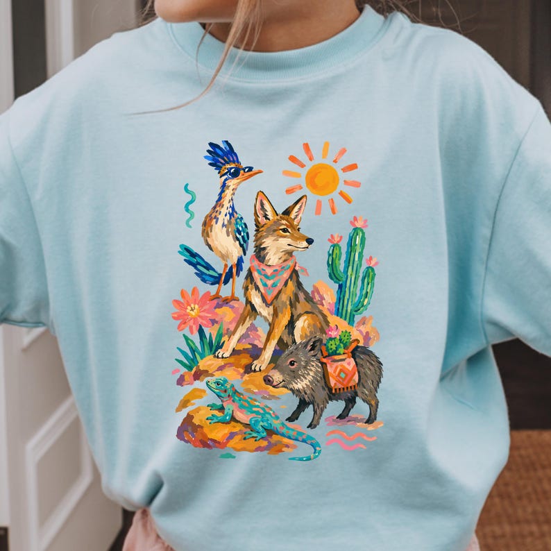 May include: A light blue crewneck sweatshirt featuring a colorful graphic print of a desert scene. The illustration includes a roadrunner wearing sunglasses, a coyote with a patterned bandana, a javelina, a lizard, a cactus, and a bright sun.