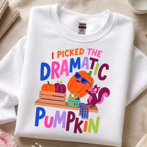 May include: A white crewneck sweatshirt featuring a colorful graphic with the text I PICKED THE DRAMATIC PUMPKIN. The design includes a large orange pumpkin wearing blue sunglasses, a purple squirrel, and smaller pumpkins in a wooden crate.