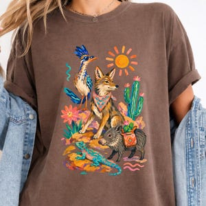 May include: A brown t-shirt featuring a colorful graphic print of a roadrunner wearing sunglasses, a coyote with a patterned bandana, a javelina carrying a small cactus, a lizard, a bright yellow sun, and a green cactus against a desert landscape.