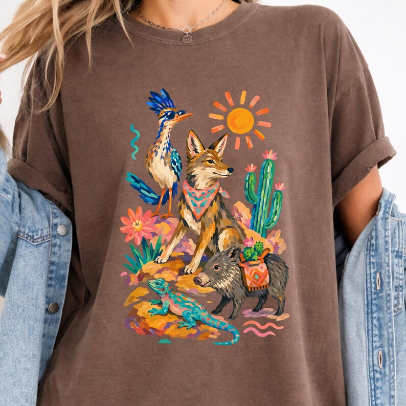 May include: A brown t-shirt featuring a colorful graphic print of a roadrunner wearing sunglasses, a coyote with a patterned bandana, a javelina carrying a small cactus, a lizard, a bright yellow sun, and a green cactus against a desert landscape.