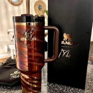 K&ouml;nnte beinhalten: Ein Stanley-Reisebecher mit einem braunen und bronzefarbenen Tigerstreifen-Design. Der Becher hat einen schwarzen Deckel und Griff. Das Stanley-Logo ist auf dem Becher und der schwarzen Box dahinter sichtbar. Eine schwarze Tasche mit dem Stanley-Logo ist ebenfalls sichtbar.