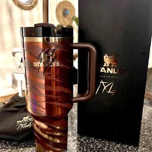 K&ouml;nnte beinhalten: Ein Stanley-Reisebecher mit braun-goldenem Tigerstreifenmuster. Der Becher hat einen Griff, einen schwarzen Deckel und einen Strohhalm. Das Stanley-Logo ist auf dem Becher und der schwarzen Box zu sehen. Ein schwarzer Beutel mit dem Stanley-Logo ist ebenfalls vorhanden.
