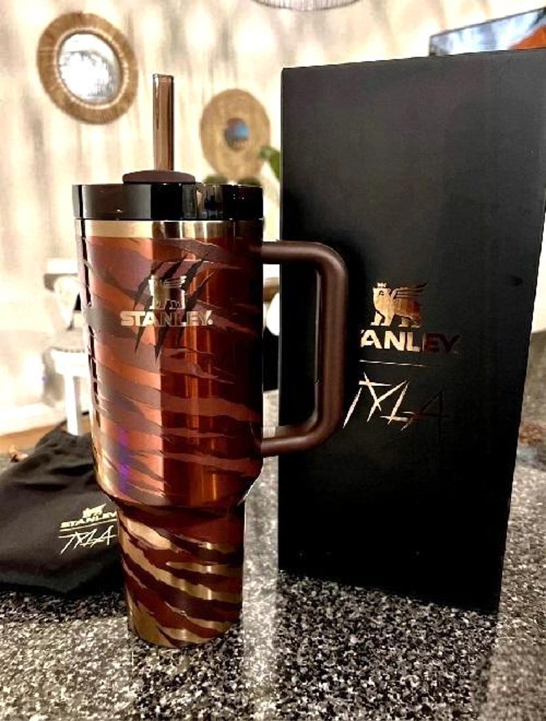 K&ouml;nnte beinhalten: Ein Stanley-Reisebecher mit braun-goldenem Tigerstreifenmuster. Der Becher hat einen Griff, einen schwarzen Deckel und einen Strohhalm. Das Stanley-Logo ist auf dem Becher und der schwarzen Box zu sehen. Ein schwarzer Beutel mit dem Stanley-Logo ist ebenfalls vorhanden.