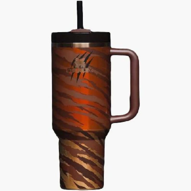 K&ouml;nnte beinhalten: Ein Edelstahl-Reisebecher mit einem Tigerstreifenmuster in Braun- und Oranget&ouml;nen. Der Becher hat einen schwarzen Deckel, einen Griff und einen schwarzen Strohhalm. Das Stanley-Logo ist auf der Seite sichtbar.