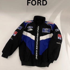 Op de afbeelding: Zwarte, blauwe en witte racejas met het woord "FORD" bovenaan. De jas heeft de tekst "FORD PERFORMANCE RACING" langs de zijkant en diverse sponsorlogo's. De jas heeft een opstaande kraag en een volledige ritssluiting.