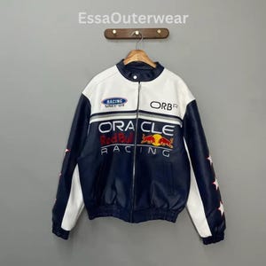 Puede incluir: Chaqueta de carreras azul marino y blanca con el texto "ORACLE Red Bull RACING" y el logotipo de Red Bull. La chaqueta tiene cremallera, cuello alzado y detalles de estrellas en las mangas. También es visible el texto "RACING SINCE '04".