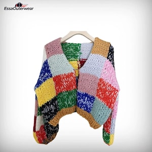 Op de afbeelding: Een kleurrijk patchwork-vest in geel, blauw, rood, roze en groen. De dikke gebreide trui hangt aan een houten hanger. Het Essa Outerwear-logo is linksboven zichtbaar. Een opvallend kledingstuk.