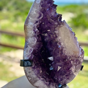 May include: A large amethyst geode specimen featuring deep purple crystalline structures and a white quartz inclusion. The geode is mounted on a black metal display stand and has a small white circular sticker with the number 8 on the top edge.