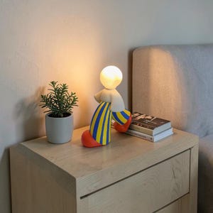 May include: A whimsical lamp shaped like a person, with a glowing bulb head. The figure has a white torso, blue and yellow striped legs, and red feet. It sits on a wooden nightstand next to a small potted plant.