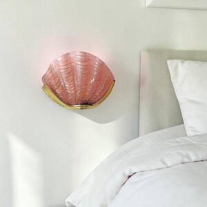 Könnte beinhalten: Eine rosa, muschelförmige Wandleuchte mit einem goldfarbenen Metallsockel. Die Leuchte ist an einer weißen Wand montiert, mit einem Bett und Kissen im Hintergrund. Die Leuchte hat ein strukturiertes, geripptes Design.