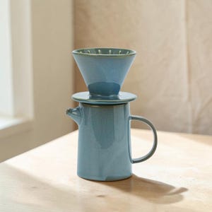 May include: A blue ceramic pour-over coffee maker with a matching filter cone. The coffee pot has a handle and a spout. The filter cone sits on top of the pot. The set is on a light-coloured wooden surface.