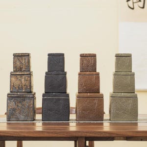 May include: Four stacks of square ceramic boxes in varying sizes and colors, including dark gray, brown, and olive green. Each box has a textured surface and a lid with a leaf design. The boxes are arranged on a wooden table.