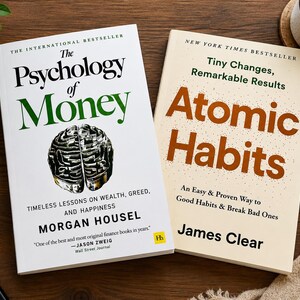 May include: Two hardcover books on a wooden surface. The book on the left is titled "The Psychology of Money" by Morgan Housel. The book on the right is titled "Atomic Habits" by James Clear.