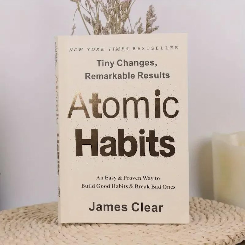May include: A hardcover book titled "Atomic Habits" by James Clear. The book has a cream-colored cover with gold lettering and the text "New York Times Bestseller." The book is on a woven surface with dried flowers.