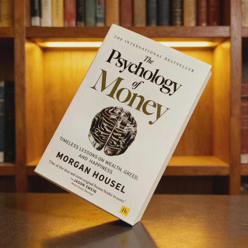 May include: A hardcover book titled "The Psychology of Money" by Morgan Housel. The cover is white with gold lettering and a graphic of a brain. The book is open, displaying the title and author against a backdrop of a wooden bookshelf.