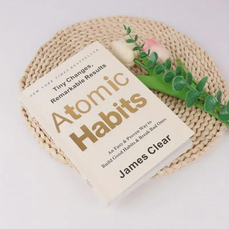 May include: A hardcover book titled "Atomic Habits" by James Clear, with gold lettering on a cream-colored cover. The book is on a woven beige placemat, with a sprig of greenery and a pink and white flower.