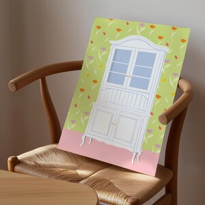 Op de afbeelding: Een print van een witte kast met blauw getinte ramen en deuren, tegen een lichtgroene achtergrond met bloemmotieven. De kast staat op een roze basis. De print staat op een houten stoel.