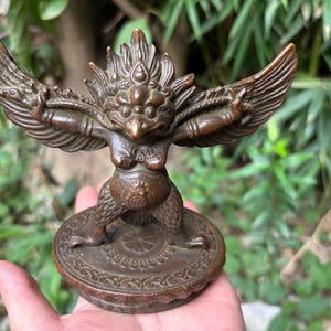 May include: A bronze-coloured statue of a mythical bird-like creature with outstretched wings, a prominent crest, and a rounded torso. The figure stands on a circular base featuring intricate, embossed geometric patterns and a central floral motif.