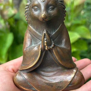 Puede incluir: Una figura de erizo color bronce en una postura sentada y meditativa con los ojos cerrados. El erizo viste una túnica de mangas anchas y mantiene sus patas delanteras juntas en posición de oración, con un pequeño rosario entre ellas.