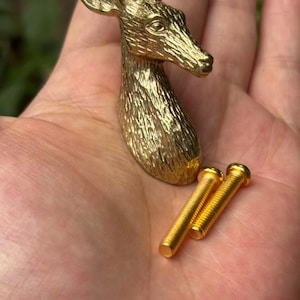 Puede incluir: Un pomo de mueble de metal dorado con forma de cabeza de ciervo y detalles de pelaje texturizado, acompañado de dos tornillos de metal dorado a juego con rosca para su instalación, con un acabado pulido y una silueta animal realista.