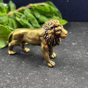 Peut inclure: Petite figurine de lion en laiton. Le lion est représenté debout, avec des détails complexes sur sa crinière et son visage. La figurine dorée est placée sur un fond sombre, avec des feuilles vertes visibles.
