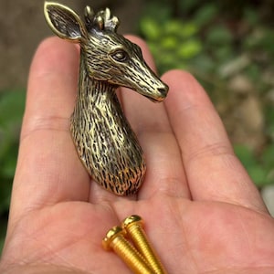 May include: A brass-toned metal cabinet knob shaped like a deer head with textured fur details and pointed ears, accompanied by two gold-colored metal mounting screws with flat heads and threaded shafts for installation on furniture or cabinetry.
