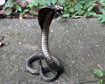 May include: A metallic cobra figurine in a striking upright defensive posture with its hood flared. The snake features a dark, weathered finish with silver highlights that accentuate the detailed scale texture across its coiled body and head.