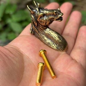 May include: A metallic, brass-toned cabinet knob shaped like a horse head with detailed mane textures, accompanied by two gold-colored metal screws with threaded shafts and flat heads for installation purposes.