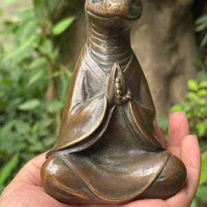 Puede incluir: Una figura de color bronce de una criatura con cabeza de serpiente en posición de meditación sentada. La figura viste una túnica con solapas plegadas y tiene las manos juntas en posición de oración, sosteniendo un pequeño rosario entre las palmas.