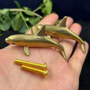 May include: Two brass dolphin-shaped cabinet knobs with a metallic gold finish, each featuring a detailed dorsal fin and tail. The set includes two matching gold-colored metal mounting screws with threaded shafts for installation on furniture.