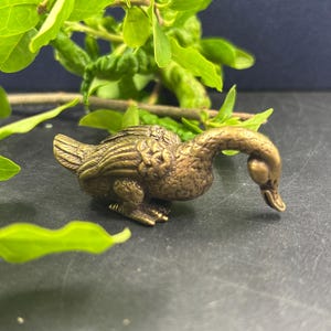 Puede incluir: Pequeña y detallada figura de pato de latón con la cabeza baja. El pato tiene plumas texturizadas y está posicionado sobre una superficie oscura, con hojas verdes en el fondo. La figura es un objeto decorativo.