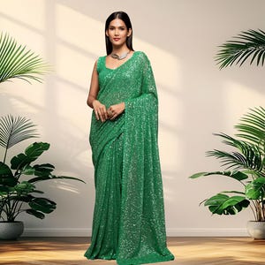 May include: A green sequined saree with a matching sleeveless blouse. The saree is draped over the body. The model is wearing a silver necklace and ring. The background is a neutral color with plants.