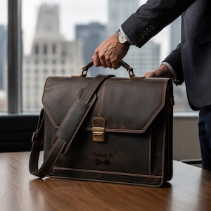 May include: A dark brown leather briefcase with a shoulder strap and a handle. The briefcase has a flap closure with a gold-colored clasp. The text "Talon K" and a bow tie graphic are visible on the front. The briefcase is held by a person wearing a gray blazer.