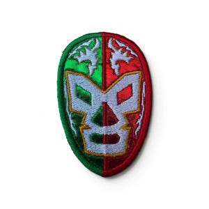 May include: An embroidered patch shaped like a luchador mask, split vertically into green and red halves. The mask features a white central design with gold outlines and white flame-like accents above the eye openings, all bordered by stitching.