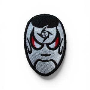 May include: An oval-shaped embroidered patch featuring a stylized character face with a light blue base, black hair, and bold black outlines. The character has sharp red eyes and a black symbol on the forehead. The entire patch has a black border.