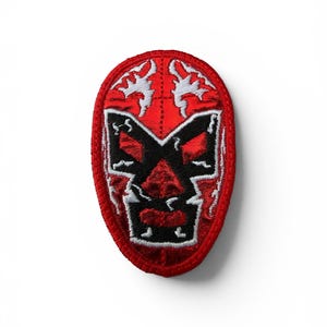 May include: An oval-shaped embroidered patch featuring a stylized red mask design. The mask has bold black outlines, white accents around the eyes and forehead, and a solid red background. The entire patch is bordered by a thick red stitched edge.