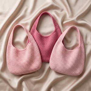 Puede incluir: Tres bolsos de ganchillo hechos a mano en diferentes tonos de rosa. Los bolsos tienen forma redondeada con una sola asa. El bolso central es de un color rosa más intenso, mientras que los otros dos son de tonos rosas más claros. Los bolsos se exhiben sobre una tela de satén.