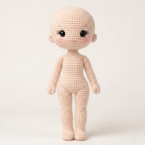Puede incluir: Una muñeca de ganchillo con tez clara. La muñeca tiene una cabeza redonda, ojos negros grandes con pestañas y mejillas rosadas. Los brazos y las piernas son proporcionados, y la muñeca está de pie.