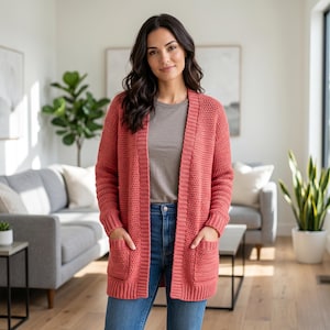 May include: A coral-colored, open-front cardigan with two front pockets. The long-sleeved sweater is made of a chunky knit material. The cardigan is knee-length and has a relaxed fit. The model is wearing a gray t-shirt and blue jeans.