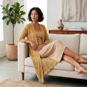 May include: A mustard yellow, open-front crochet cardigan with a floral pattern. The long cardigan is knee-length and has three-quarter sleeves. The woman is wearing a beige dress and sandals.