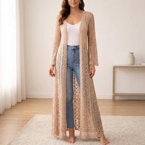 May include: A long, open-front, beige crochet cardigan. The cardigan has long sleeves and a floor-length design. The crochet pattern is intricate, creating a delicate, airy look. The cardigan is worn over a white top and blue jeans.