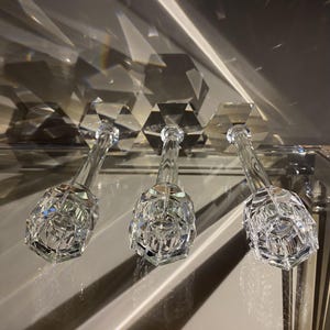 K&ouml;nnte beinhalten: Drei Kerzenhalter aus klarem Glas mit facettiertem, sechseckigem Design. Jeder Halter verf&uuml;gt &uuml;ber einen hohen, schlanken Stiel und einen breiten, geometrischen Fu&szlig;, der das Licht reflektiert und scharfe Schatten sowie helle Lichtpunkte erzeugt.